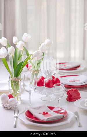 Happy easter. Decor and table setting of the Easter table is a vase with white tulips and dishes of red and white color. Easter colored eggs with whit Stock Photo