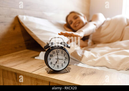 Young beautiful blonde woman lying in bed in morning. Positive happy model reaching alarm clock with hand. . Daylight. Good mood Stock Photo