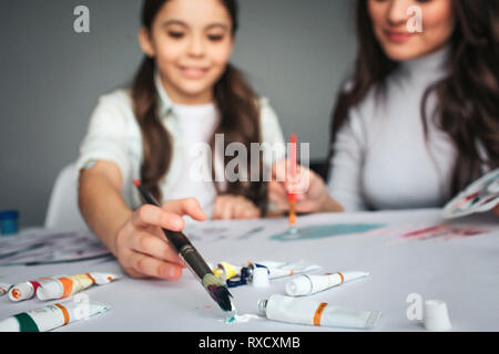 Beautiful brunette caucasian mother and daughter paint together in room. Cut view and blurred picture. Happy girl put brush into color. Mother sit Stock Photo