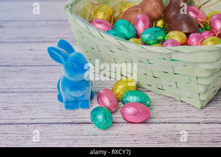 blue easter bunny beside colored chocolate eggs, in the background an easter basket on white wooden background Stock Photo