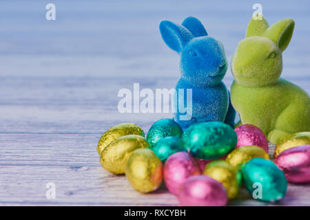 blue easter bunny and green easter bunny with wrapped chocolate eggs against white wooden background with copy space Stock Photo