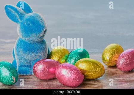 blue easter bunny with wrapped chocolate eggs against white wooden background with copy space Stock Photo