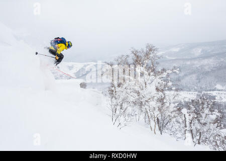 Backcountry skier jumps off a cornice near the summit of a small peak in backcountry Hokkaido, Japan. Yellow jacket, blue backpack powder skiing in Ja Stock Photo