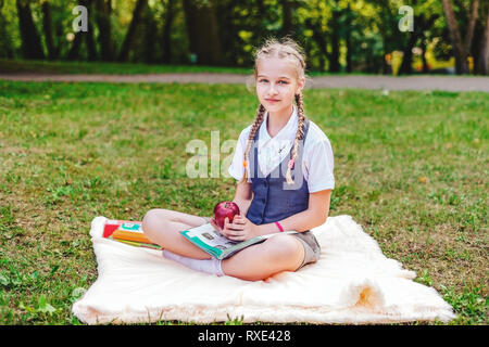 portrait of schoolgirl teenager with pigtails sitting in park on coverlet with apple and books Stock Photo