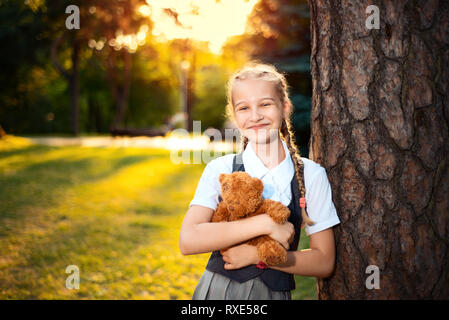 portrait of schoolgirl with pigtails in uniform at sunset. the student stands near a tree and holds a soft bear toy Stock Photo