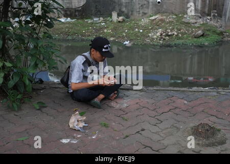 A man hold a smartphone while sitting on a dirty pavement yard beside a river in Jakarta, Indonesia. Stock Photo