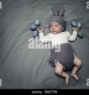 Cute little baby dressed like an Easter bunny, adorable sleeping kid on the gray blanket background with stylish decorated gray Easter eggs, happy rel Stock Photo