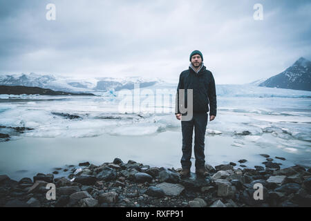 Young man standing in front of Fjallsarlon iceberg lagoon at the south end of the glacier Vatnajokull, with floating icebergs that calve from the edge Stock Photo