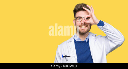 Young handsome scientist man wearing glasses doing ok gesture with hand smiling, eye looking through fingers with happy face. Stock Photo
