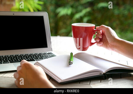 Work concept: One hand on red mug and another on table with laptop and note pad in view Stock Photo