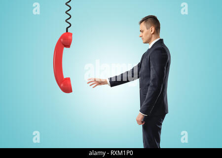 Side view of businessman standing and reaching out for big red landline phone receiver dangling down on wire from above. Stock Photo