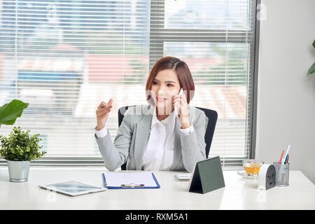 Cheerful asian female talking on mobile phone while sitting on desk with laptop. Business woman in casuals making a phone call and laughing. Stock Photo