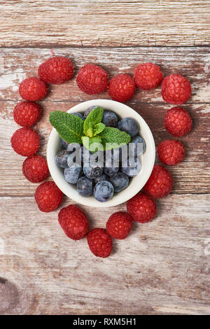 raspberries on wooden surface in a heart shape, there are blueberries in a white bowl in the middle of the heart Stock Photo