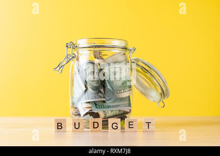 glass jar filled with dollars and word budget made of wooden blocks on yellow background Stock Photo