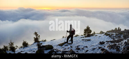 Back view of tourist hiker with backpack and trekking sticks on rocky mountain peak enjoying panoramic view of valley covered with clouds stretching t Stock Photo