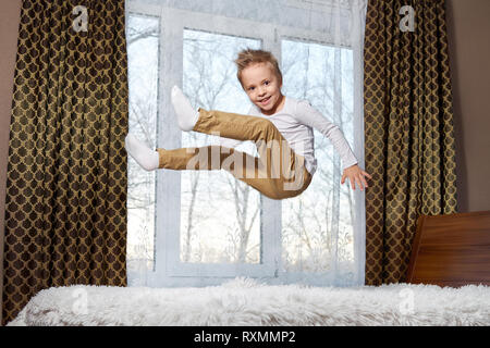 child fun home. Beautiful blond child 6 years old fooling around morning to room. Kid smiling jumping to bedroom on bed. Stock Photo