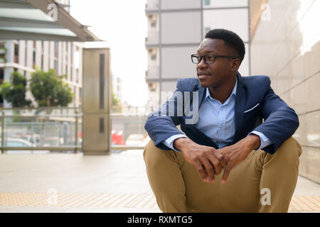 Young thoughtful African businessman sitting outside the subway train station Stock Photo