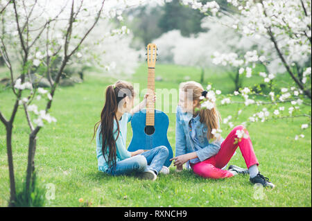 two happy friends young girls sitting on green grass together looking at custom guitar under blossoming trees in spring May park Stock Photo