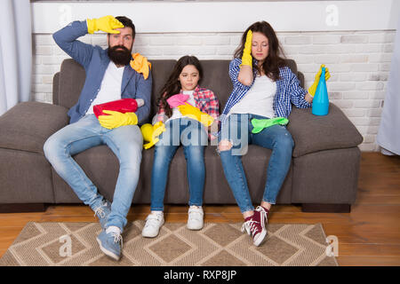 Tired parents and kid. Cleaning all day exhausting occupation. Exhausting cleaning day. Family mom dad and daughter with cleaning supplies sit on couch. Family care about cleanliness. Finish cleaning. Stock Photo