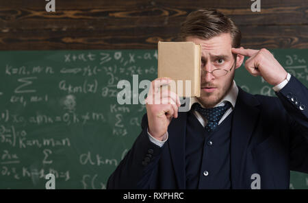Closeup portrait of student holding book in front of his face and pressing finger over forehead. Thoughtful intelligent man in classroom Stock Photo