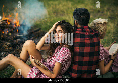 Smiling brunette with braces leaning on her friend. Turn back guy looking at fire while two beautiful girls read books. Relaxation in nature Stock Photo