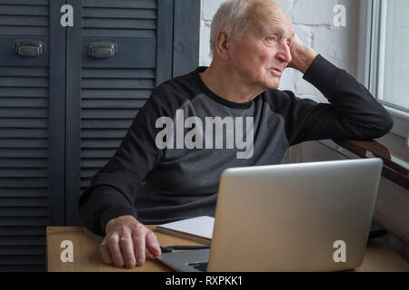 An elderly man looks out the window in front of an open laptop, counting taxes, selective focus, free space for text. Stock Photo