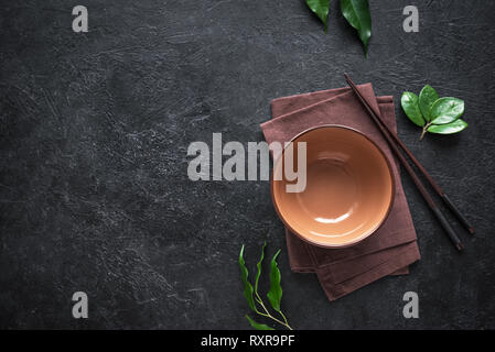 Ceramic bowl and chopsticks on black table, top view, copy space. Empty bowl of Chinese noodles, plant leaves and chopsticks, asian chinese japanese t Stock Photo