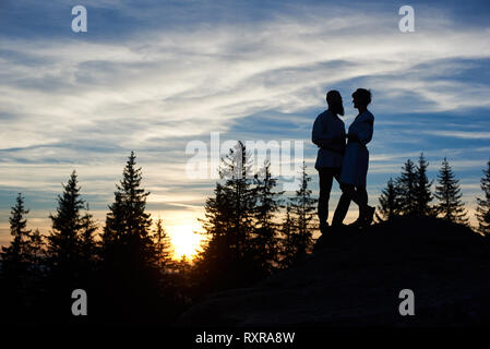 Silhouettes of young couple in love standing on the top of mountain in the evening. Bearded man and happy girl enjoying beautiful sunset, looking to e Stock Photo