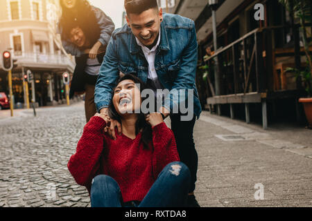 Excited young woman being pushed on skateboard by her boyfriend outdoors on street, with friends piggybacking in background on the street. Group of fr Stock Photo