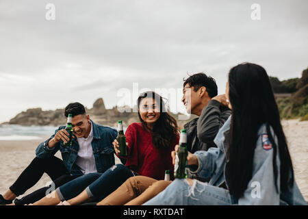 Cheerful young asian men and women sitting at the beach having beers. Two couple partying on the beach. Stock Photo