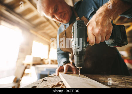 Senior male carpenter drilling a hole in wooden plank on workbench. Focus on carpenter hand preparing furniture. Stock Photo