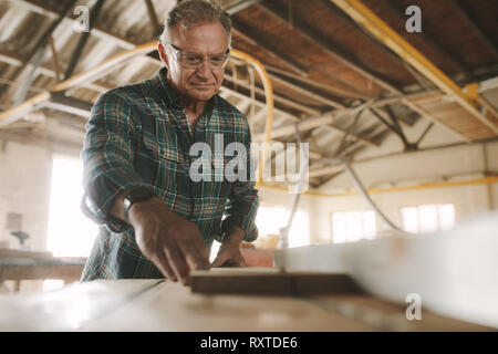 Senior male carpenter working on electric table saw machine cutting wood planks. Skilled mature carpenter working in carpentry workshop. Stock Photo