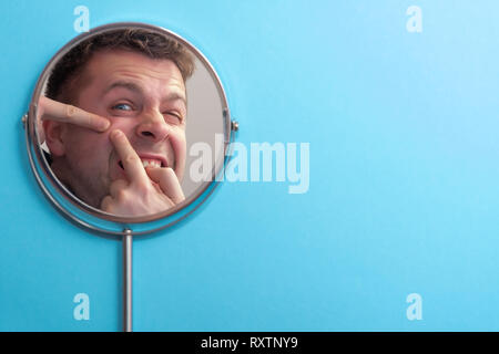 Young man touching his face and crushing pimple in mirror on blue background Stock Photo