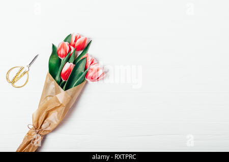 View from above bouquet of fresh red tulip flowers in brown kraft paper next to golden scissors on white wooden background, copy space. Stock Photo