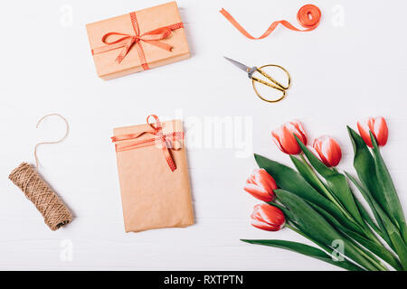 Festive flat lay arrangement of process of wrapping gifts for holidays. Box with bow next to scissors, jute, red ribbons and bouquet of freshly cut tu Stock Photo