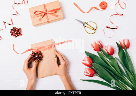 Woman's hands decorating box with red bow near scissors, ribbons and bouquet of tulip flowers on white wooden table, top view. Festive flat lay compos Stock Photo