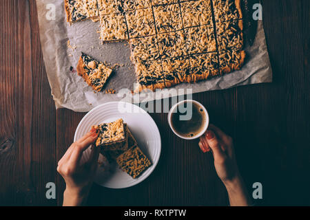 Woman's hands holding cookies and cup of coffee next to baking pan of hot homemade cakes on brown wooden table, top view. Stock Photo