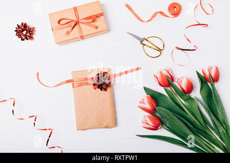 Festive flat lay composition of process of packing gifts for holidays. Box with bow next to scissors, red ribbons and bouquet of freshly cut tulip flo Stock Photo
