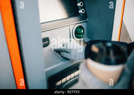 Close-up of young woman's hands holding coffee and entering PIN on ATM keypad on city street in winter day. Stock Photo
