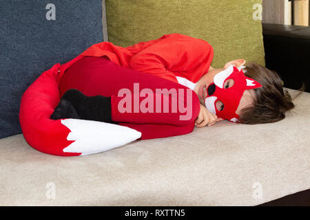 Child in fox fancy dress mask lying curled up on sofa imitating a real fox Stock Photo