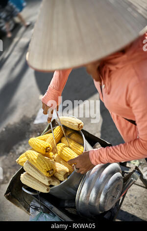 Street seller in a asian conical hat sells the boiled yellow corn in the metal saucepan on the sunny background outdoors. She takes one with the help  Stock Photo