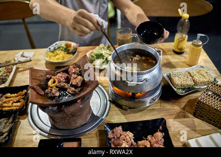 Man is sitting by the wooden table and cooking soup on the hot pot in the local traditional cafe in Vietnam. Varied food on the plates and bowls are a Stock Photo