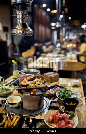 Fresh fish steak and shrimps are cooking on the hot pot with burning coal on the wooden table in the vietnamese cafe. Varied food on plates are around Stock Photo