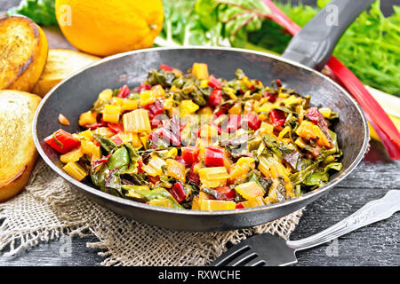 Warm chard salad with orange and onion in a frying pan on sackcloth, bread, fork on a dark wooden board background Stock Photo