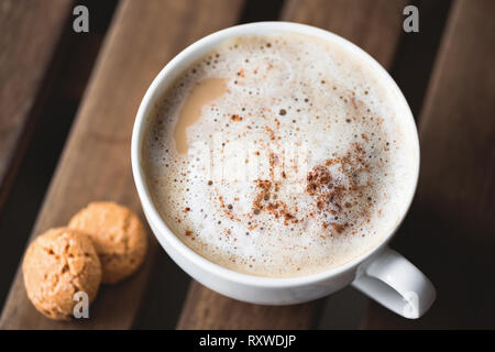 Cappucino with cinnamon in cup served with few amaretti cookies on wooden table. Breakfast in cafe Stock Photo