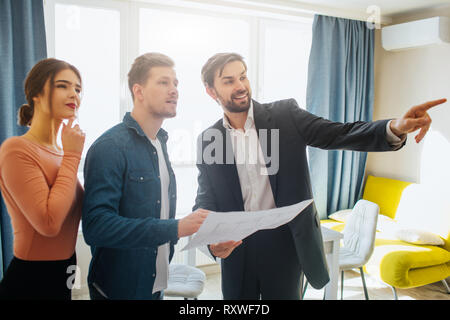 Couple buy or rent apartment together. Realtor point forward. Couple look at that way. Positive and thoughtful. Business talk. Holding apartment plan. Stock Photo