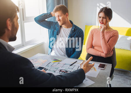 Couple buy or rent apartment together. Confused suspicious young man and woman have doubts. They sit in front of realtor. He reach hand and point Stock Photo