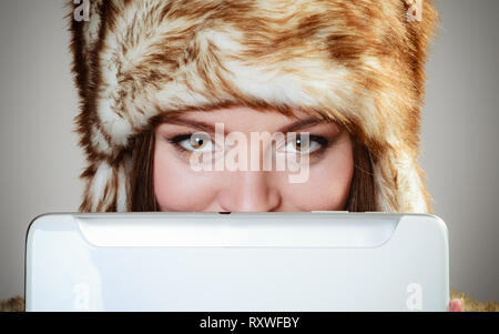 Technology internet concept. Closeup fashion woman in winter clothes fur cap covering her face with tablet computer studio shot on gray Stock Photo