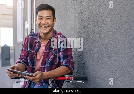 Happy young man leaning on his bicycle using a tablet Stock Photo