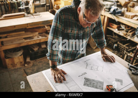 Senior carpenter studying blueprint at his workshop before starting his work. Carpenter checking drawing in carpentry workshop. Stock Photo
